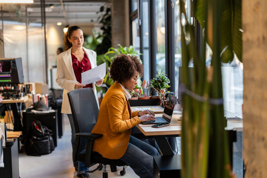 Businesswoman working at modern office desk while colleague stands nearby