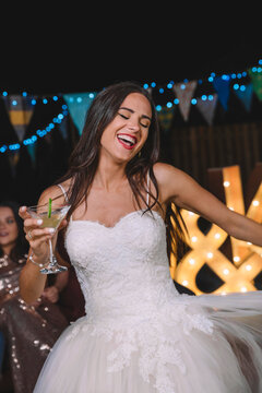 Happy bride laughing and dancing while holding cocktail on an outdoor night party