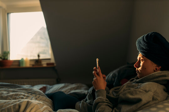 Young woman with head wrapped in a towel lying in bed at home using smartphone