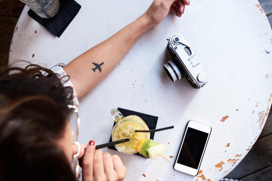 Young woman with airplane tattoo on her forearm sitting in a coffee shop drinking pineapple smoothie