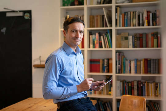 Portrait of confident businessman with tablet leaning at table with bookshelf in background
