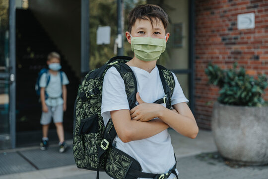 Little boy wearing protective face mask with arms crossed standing in front of school building