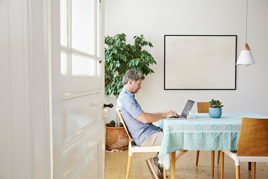 Mature man at home sitting at table, using laptop