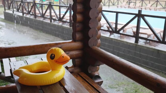 An inflatable duck lies on wooden table in gazebo at recreation center against the backdrop of pool covered with rain