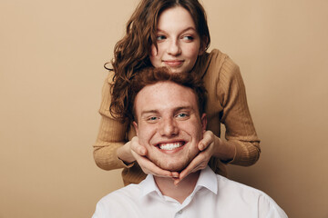 Fototapeta premium Smiling young couple with woman holding man’s face gently. Warm beige background, casual clothing. Concept of love, affection, happiness, and close relationship between partners.