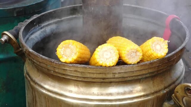 Corn on the cob is cooking in a metal pot on a street cart. People move around the market. The sun shines bright in the afternoon sky while steam rises from the pot.