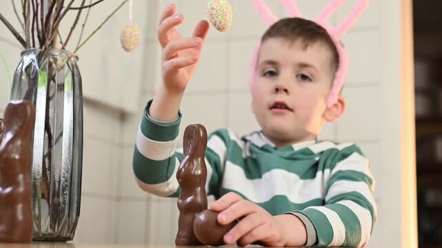 A preschool boy in a bunny costume eats a chocolate egg and a chocolate bunny at home in the kitchen, celebrating Easter. Boy playing with chocolate easter rabbit