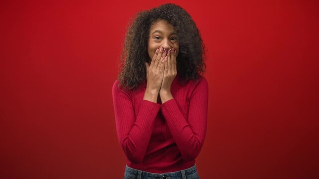 Woman covers mouth with hands in studio with red backdrop and red sweater, curly hair and denim jeans; surprise delight.