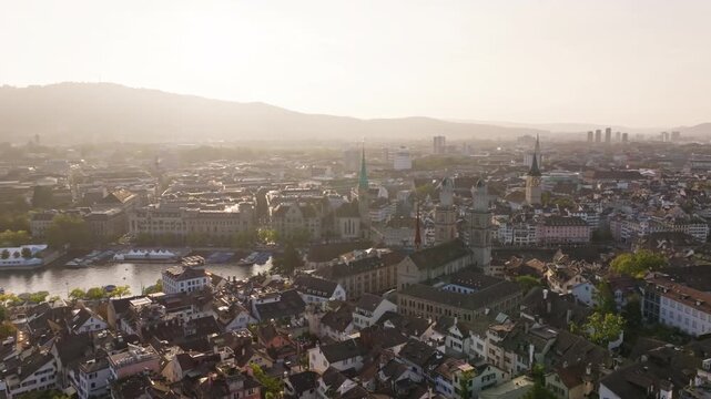 Cinematic drone shot at dawn orbiting around the Grossm&uuml;nster with sweeping views over the Limmat and Quaibr&uuml;cke in Zurich. Warm golden hour light, calm river reflections, and iconic Swiss city skylin