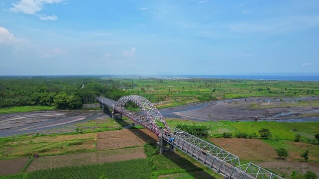 Aerial drone footage of the metallic bridge of Pandanwangi, with a vast green agricultural and grass valley behind, with black stone dry river, blue sea, in Lumajang regency, Java island, Indonesia