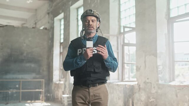 Handsome Caucasian journalist reporting live in war zone. Brave man wearing helmet and bulletproof vest speaking with microphone at camera in abandoned building. Reporting news for TV media.