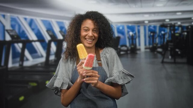 Woman with curly hair holding two colorful popsicles in her hands and smiling with eyes closed, apron visible inside a building gym with treadmills; carefree joy.