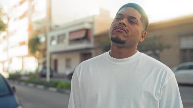 Man with beard tilts head up and looks skyward on a sunny street wearing white shirt, subtle smile emerges; serenity.