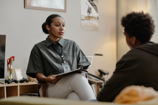 Black young adult woman social worker holding clipboard engaging in conversation with teenager indoors, both seated and facing each other during counseling session