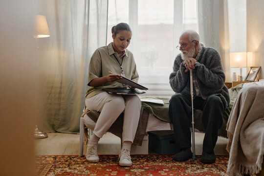 Young adult Black woman social worker sitting on bed holding clipboard, talking with senior Caucasian man with beard using cane in residential room during consultation session