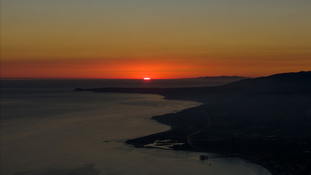 Aerial view of the sun sinking below the horizon, casting a fiery glow over the tranquil waters and rugged coastline, Malibu, California, United States.