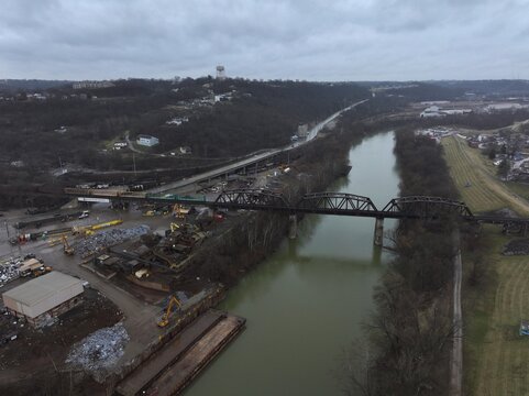Aerial view of the murky river reflecting the overcast sky, a dark metal bridge spanning its width near industrial activity, Newport, Kentucky, United States.