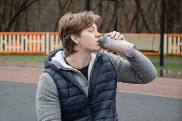 Fototapeta premium Young man sits on an outdoor gym bench holding protein shake, wearing casual vest. He relaxed, enjoying moment of rest during workout. Recovery after workout.