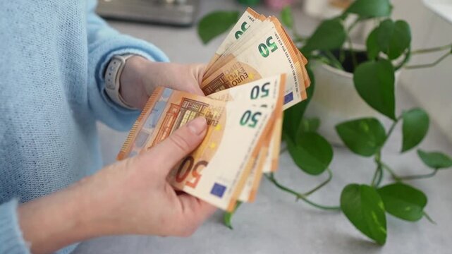 Hands counting euro banknotes on countertop, soft natural light, green pothos plant, blue knit sweater, meticulous thumb through notes, planning savings, personal finance mood Hands counting euro bank