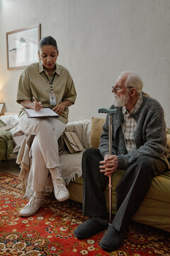 Young adult Black woman social worker sitting on couch writing on clipboard while talking with senior Caucasian man holding cane, both engaged in conversation in home setting
