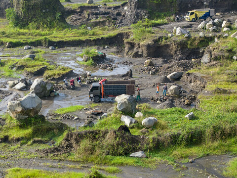 Aerial view of trucks and workers amidst a rugged, rocky landscape with patches of green vegetation, Kali woro, Klaten Regency, Jawa Tengah, Indonesia.