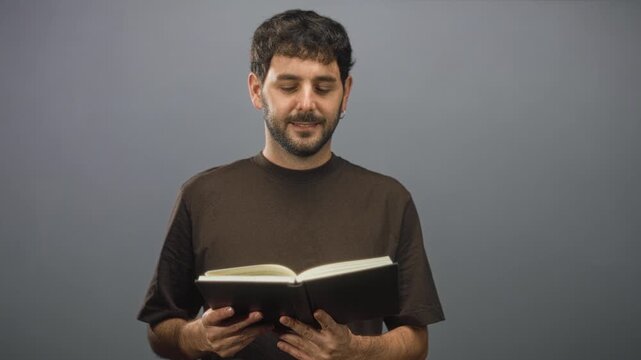 Young hispanic man with beard holding open book, hands visible, slight smile, earring visible, reading in studio; contemplation.
