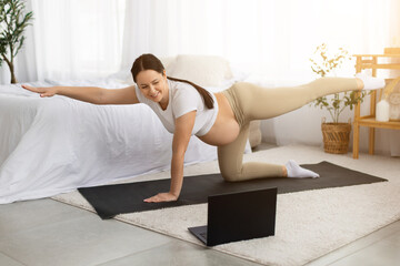 Obraz premium Pregnant young woman performing prenatal balance yoga pose at home while following online workout on laptop. Healthy maternity exercise and pregnancy fitness routine