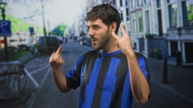 Man in blue and black football jersey pointing both index fingers upward on a street with buildings and bicycles and parked cars; team pride.