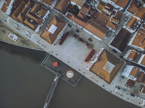 Aerial view of terracotta rooftops meeting the dark river edge, framing the light square of Praca Gomes Freire, Alcacer do Sal, Setubal, Portugal.