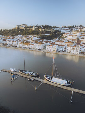 Aerial view of a serene river reflecting the bright sky, with boats docked at a wooden pier against a backdrop of white buildings and a hilltop castle, Alcacer do Sal, Setubal, Portugal.