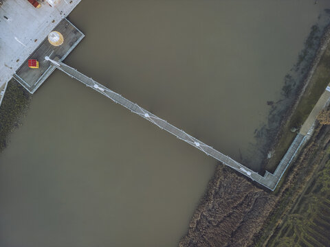 Aerial view of a narrow bridge stretching across the muddy river, connecting to a concrete platform, contrasting with the natural textures of riverbank vegetation, Alcacer do Sal, Setubal, Portugal.