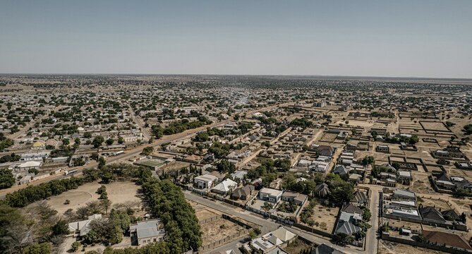 Aerial view of a sprawling townscape of tightly packed buildings interspersed with green trees, under a pale sky, Daura, Katsina, Nigeria.
