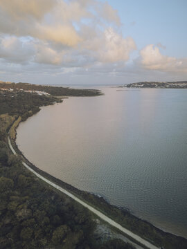 Aerial view of a serene lagoon reflecting the soft, diffused light of the sky, bordered by a dark, lush forest and a sandy path, Lagoa de Obidos, Leiria, Portugal.