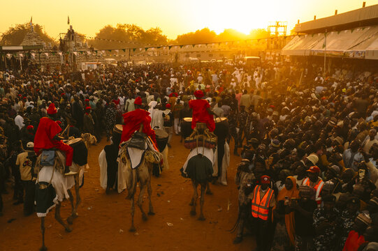 Argungu, Nigeria - 13 February 2026: Aerial view of a vibrant crowd gathered under the golden sunset at the Argungu Fishing Festival, with camel riders in striking red attire leading the way.