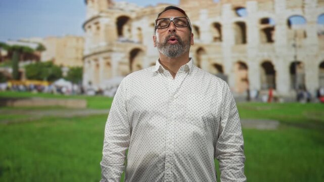 Man with beard and glasses, eyes closed and head tilted back at building coliseum; serenity and reflection.