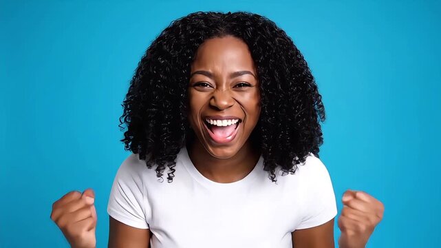 Excited black woman shouting with joy and clenching fists for success celebration on bright blue studio background wearing a white casual t shirt