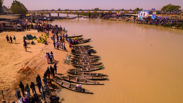 Argungu, Nigeria - 13 February 2026: Aerial view of the sun-drenched Matan Fada River, where dark wooden boats contrast against the ochre water, framed by crowds gathered on the banks and the bridge.