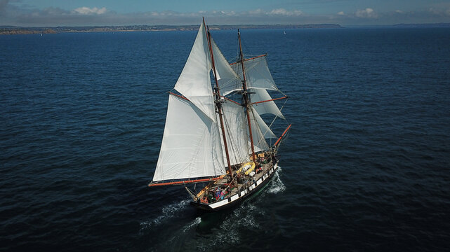 Plougonvelin, France - 12 August 2021: Aerial view of a majestic sailboat, its white sails billowing against the deep blue sea, slicing through the water leaving a frothy wake.