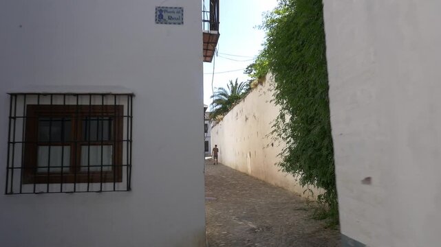 Wide tracking shot following a person walking down the narrow, cobblestone Calle del Rosal. Features whitewashed walls with green climbing plants, traditional architecture, and bright morning sun.