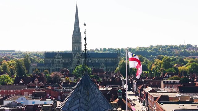 Drone aerial view of Salisbury Cathedral rising above the historic city of Salisbury in Wiltshire, England. The cathedral spire dominates the skyline while an English flag waves above the rooftops. Sc