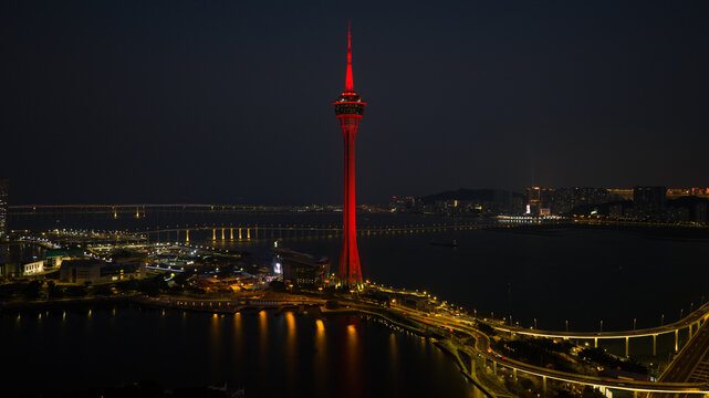 Aerial view of the Macau Tower piercing the night sky, its red glow reflecting on the waters below and illuminated city lights, Macau, Macao.