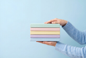 Woman hands in a blue sweater hold a stack of pastel colored books against a light blue background