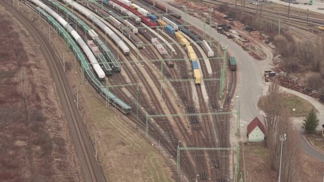 Aerial drone view of multiple freight trains standing on curved railway tracks in a large cargo yard. Colorful shipping containers and tank cars are arranged along the rail lines.