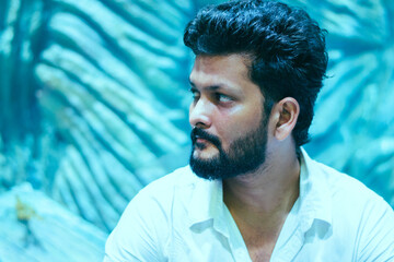 A focused young Indian man with a beard looks away in a close-up profile portrait against a blurred, vibrant teal background indoors. © DiversePixels