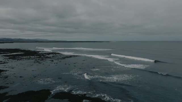 Aerial drone view of a surfer riding a clean ocean wave along the rugged coastline of Iceland under an overcast sky. Cold water surfing, adventure travel and dramatic Nordic landscape concept.