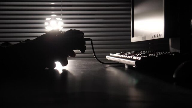 Person's silhouetted hands holding a wired gamepad while playing computer video games at a computer desk in a dark room, with light shining through window blinds
