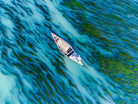 Aerial view of a solitary boat glides across the shimmering, algae-streaked waters, creating a mesmerizing dance of light and shadow, Rajshahi, Rajshahi Division, Bangladesh.