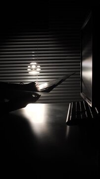 Vertical video person's hand and arm appearing as a silhouette, reaching for a computer keyboard in a dimly lit office space at night, with light filtering through horizontal blinds