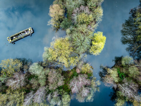 Aerial view of a boat gliding past islands, a serene contrast of blue water against the vibrant foliage, creating a tranquil scene on the River Thames, Reading, Berkshire, United Kingdom.