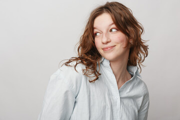 Obraz premium Young woman with curly brown hair wearing a white shirt looks sideways with a slight smile on a light background. Concept of natural beauty, casual style, and positive mood.
